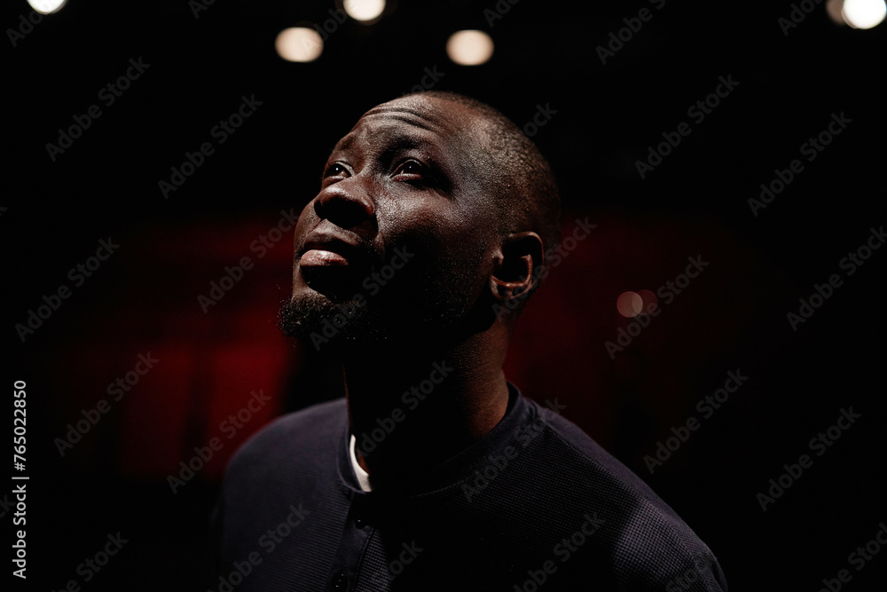 Dramatic portrait of Black adult man looking up into spotlight while standing on stage in theater with low light copy space