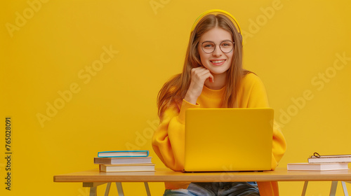 Full length portrait of a smiling student girl sitting at the table with laptop and books isolated over on yellow color background professional photography.