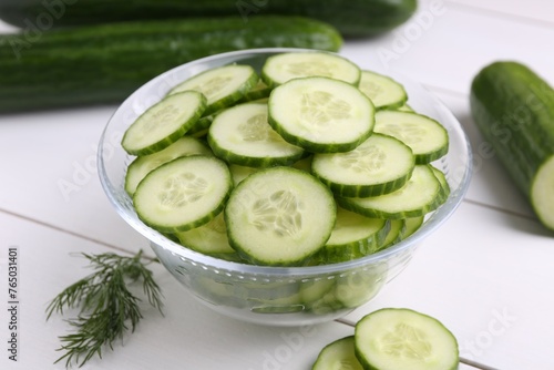 Cut cucumber in glass bowl, fresh vegetables and dill on white wooden table, closeup