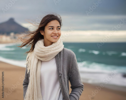 A woman wearing a white scarf and a gray sweater stands on a beach