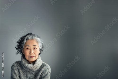 femme retraitée asiatique, origine japonaise, avec les cheveux gris, blancs et noirs, poivre et sel, avec un pull gris à col roulé face objectif, maquillée sur fond gris avec espace négatif copy space photo