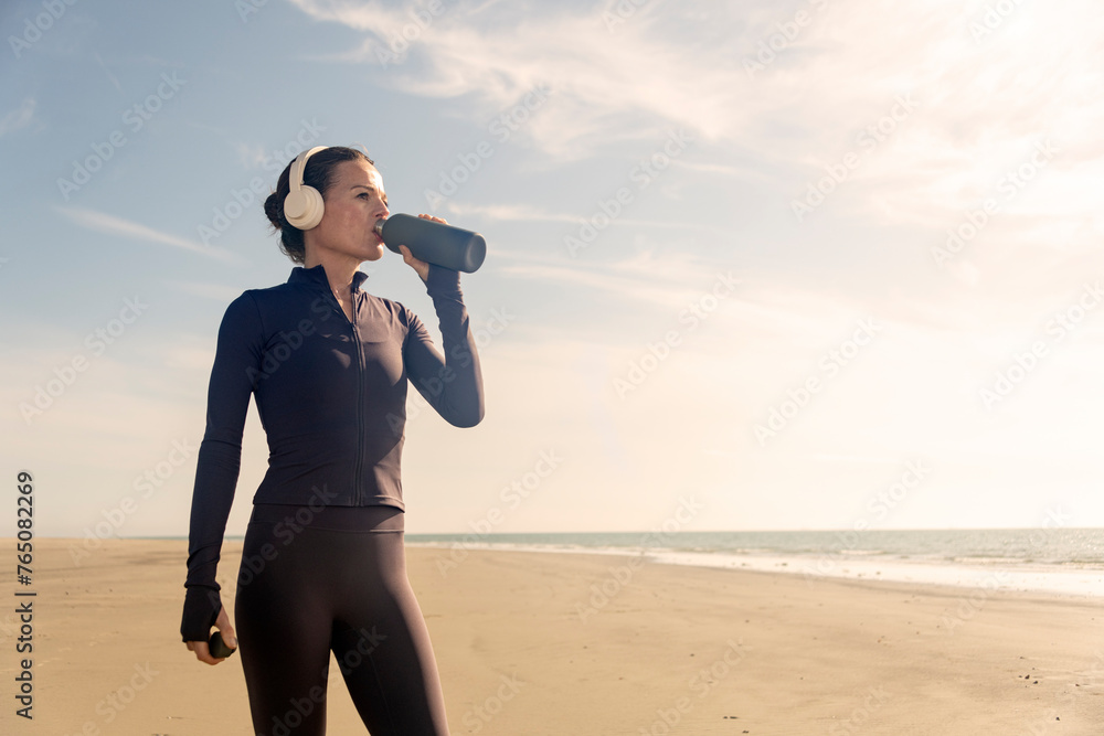 Obraz premium Sporty woman taking a drink of water from a bottle, exercise outside at a beach