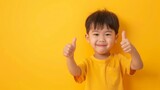 Cheerful Asian Toddler Giving Thumbs Up on Solid Yellow Background. Copy space.