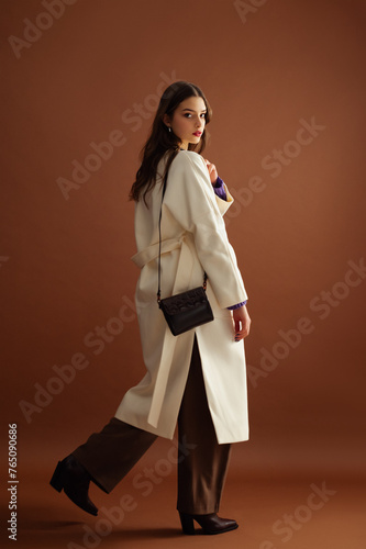 Canvastavla Fashionable confident woman wearing white coat, trousers, leather pointed toe boots, with small stylish shoulder bag, posing on brown background