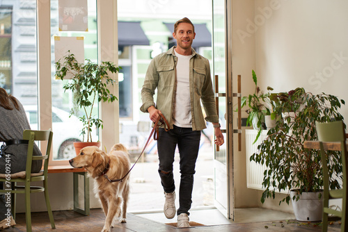 Portrait of handsome young man walks into the cafe with his dog on a leash, enters pet-friendly coffee shop, opens the door