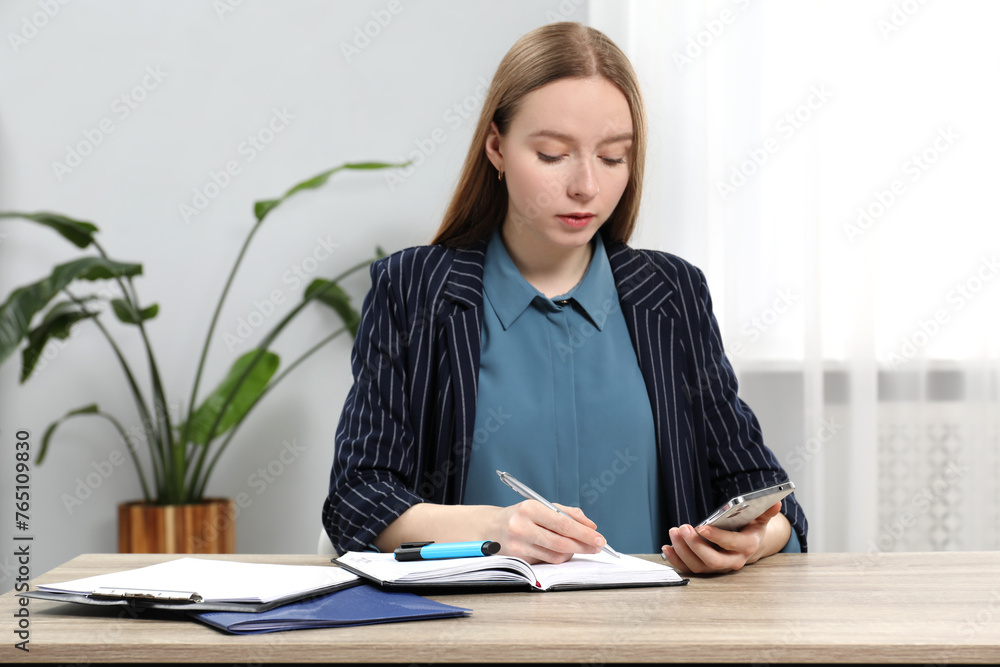 Woman taking notes while using smartphone at wooden table in office Woman taking notes while using smartphone at wooden table in office