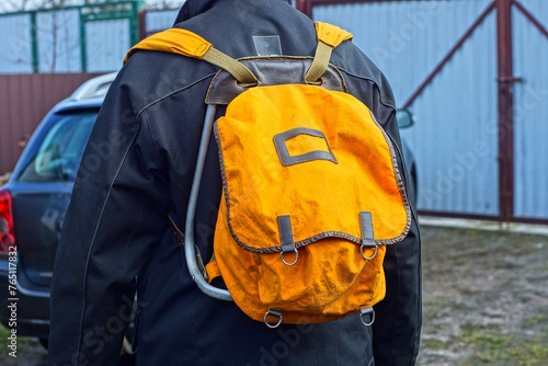 male old old-fashioned small closed yellow backpack with fabric backpack behind the back of a tourist dressed in a black jacket near a gray iron fence on the street during the day