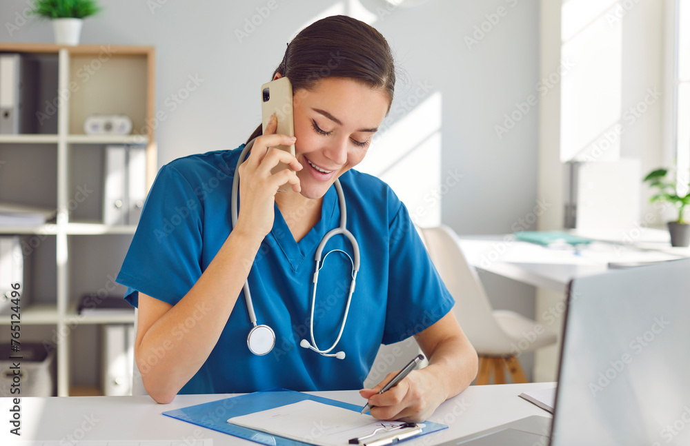 Friendly young woman doctor, nurse, reception specialist in blue scrubs with stethoscope sitting in office workplace, making phone calls, talking to customers, scheduling appointments and taking notes Friendly young woman doctor, nurse, reception specialist in blue scrubs with stethoscope sitting in office workplace, making phone calls, talking to customers, scheduling appointments and taking notes