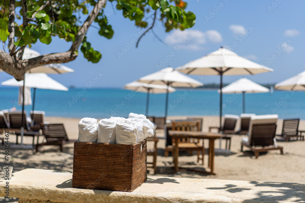 Roll up of white towels in basket on white table with copy space on blurred beach sunbeds and white umbrella background, resort vacation