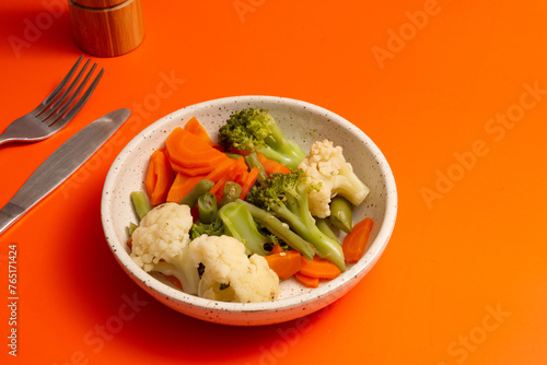 Steamed cauliflower, carrots, brocolli and vegetables inside a ceramic bowl ina orange background in front view photo