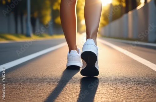 Female legs in white sneakers run along an asphalt path in the setting sun  bottom view