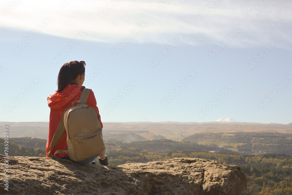 A girl tourist with a backpack sits on the top of a mountain and enjoys the beautiful views. A walking woman in an orange jacket rests on a rock, looking at the beautiful sunny landscape of the