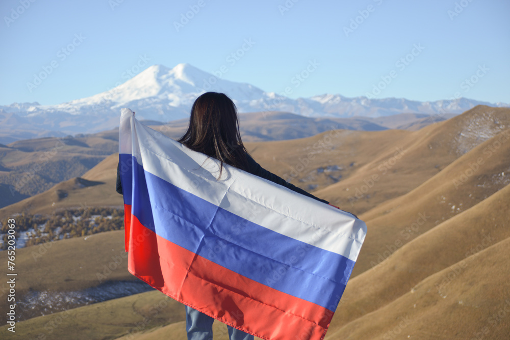 A young brunette woman stands against the backdrop of the snow-capped Mount Elbrus, looking at the beauty of the mountains, holding a Russian flag in her hands. Tricolor against the backdrop of snow
