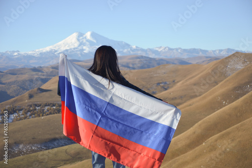 Obraz na płótnie A young brunette woman stands against the backdrop of the snow-capped Mount Elbrus, looking at the beauty of the mountains, holding a Russian flag in her hands. Tricolor against the backdrop of snow w salonie