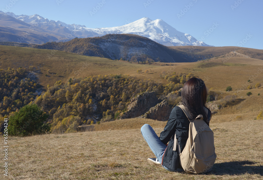 A female traveler with a backpack behind her sits on the grass and enjoys the views of Elbrus. View of Elbrus, North Caucasus, Russia.