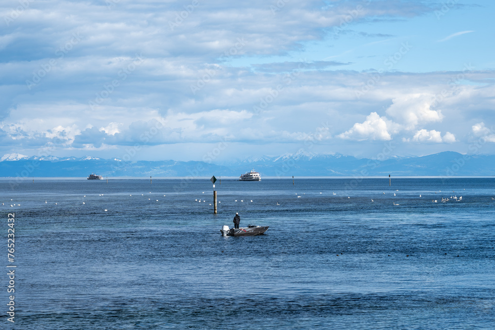 Fototapeta premium Angler auf Boot genießt den Blick über den See mit Schwänen, Dampfern und dem Alpenpanorama im Hintergrund. Konstanz, Bodensee, Baden-Württemberg, Deutschland, Europa.
