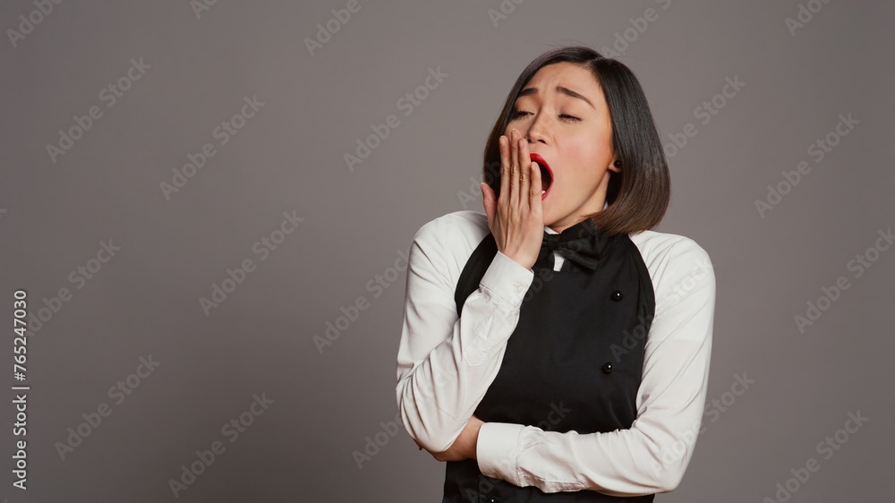 Exhausted waitress with apron yawning over grey background, feeling overworked and dealing with burnout after multiple restaurant events. Asian catering worker feeling tired in studio. Camera B. Exhausted waitress with apron yawning over grey background, feeling overworked and dealing with burnout after multiple restaurant events. Asian catering worker feeling tired in studio. Camera B.