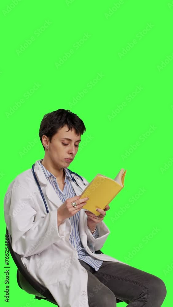 Side view Medic specialist reading a medicine literature book sitting on a chair, enjoying healthcare industry lecture against greenscreen backdrop. Doctor in white coat reads for relaxation. Camera B Side view Medic specialist reading a medicine literature book sitting on a chair, enjoying healthcare industry lecture against greenscreen backdrop. Doctor in white coat reads for relaxation. Camera B
