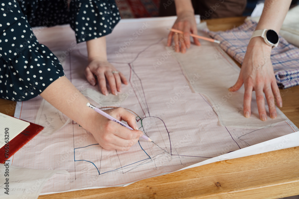 Two women examining and discussing dress pattern drawings on a wooden table, with a focus on the creative process. Two women examining and discussing dress pattern drawings on a wooden table, with a focus on the creative process.