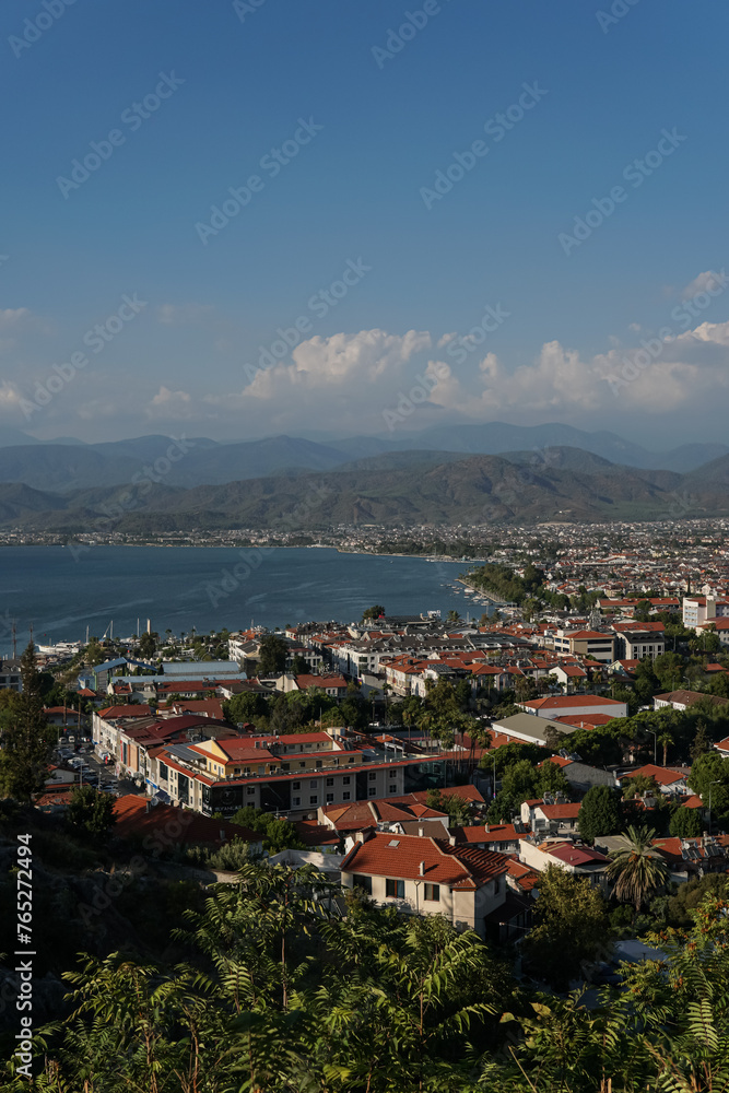 A view of the city Fethiye in Turkey, a clear blue sky, the sea and mountains, a large marina on one side,  buildings have red roofs, and greenery around the town