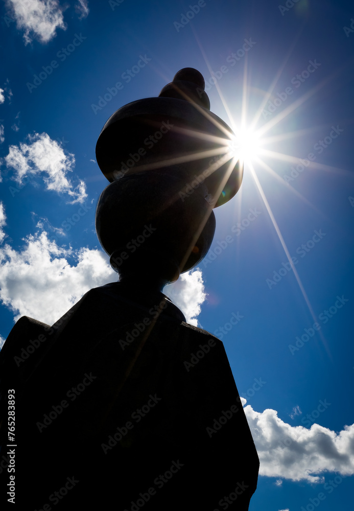 A dark silhouette of the top of an ornate tombstone stands out against a vibrant blue sky with fluffy white clouds and a bright sunburst.