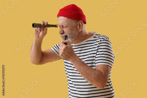 Mature sailor with smoking pipe looking through spyglass on yellow background