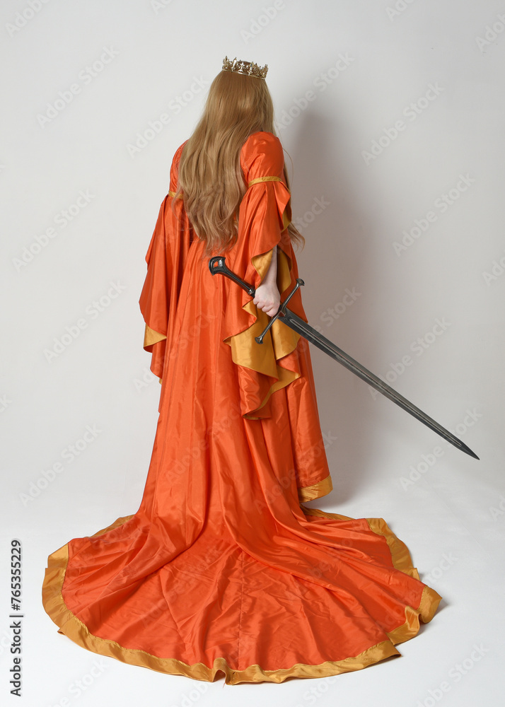 Obraz premium Full length portrait of plus size blonde woman, wearing historical medieval fantasy gown, golden crown royal queen. Standing pose backview, holding sword weapon, isolated studio background.