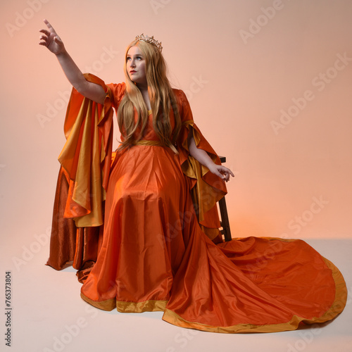 Full length portrait of plus size blonde woman, wearing historical medieval fantasy gown, golden crown royal queen. sitting pose on throne chair, isolated studio background.