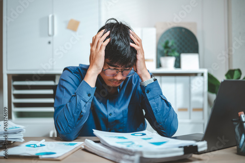 depressed, document, frustration, loss, nervous, overworked, headache, paperwork, stress, fail. A man is sitting at a desk with a laptop and papers in front of him. and he is in a state of distress.