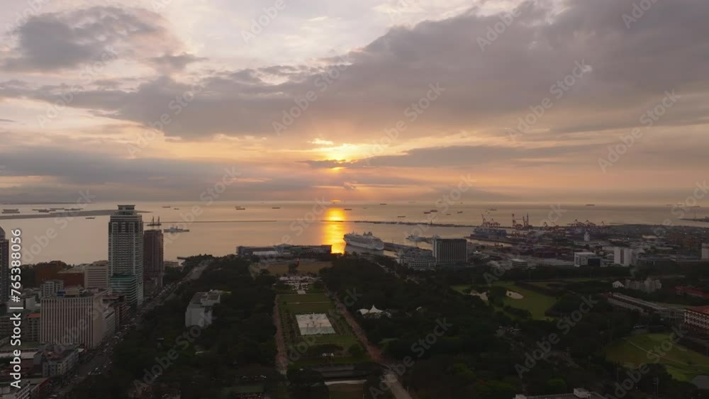 Fly above city park at sea coast. High rise buildings in city and huge cranes in harbor against colorful sunset sky. Manila, Philippines Fly above city park at sea coast. High rise buildings in city and huge cranes in harbor against colorful sunset sky. Manila, Philippines