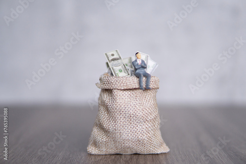 Closeup image of miniature businessman sitting on a large sack overflowing with U.S. dollar bills. Copy space for text. Success, profit, capitalist concept.
