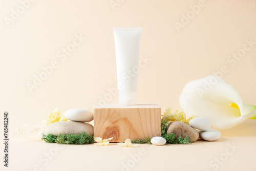 White tube of cosmetic cream with wood podium and flowers, moss, stone on pastel beige background. Close up, copy space