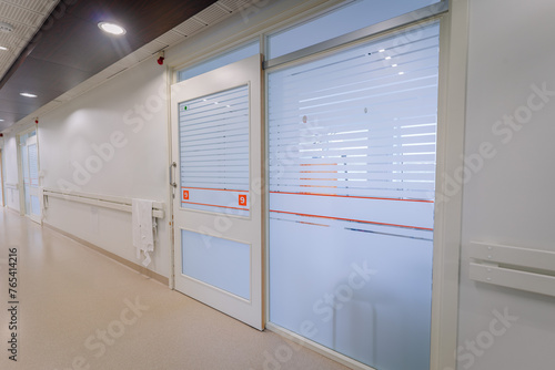 a bright hospital corridor with numbered doors, frosted glass windows, handrails, and a paper towel dispenser. photo