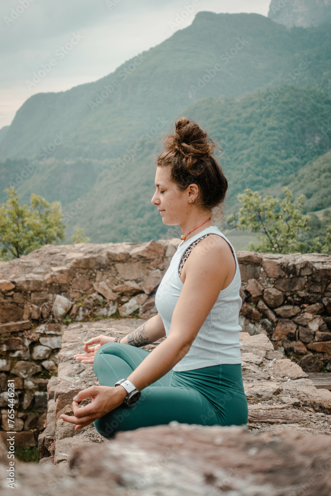 Naklejka premium A Woman Achieves Inner Peace through Yoga and Meditation in Lotus Seat, Surrounded by the Tranquil Ambiance of an Old Stone Wall amidst the Lush Greenery of a Forest Backdrop