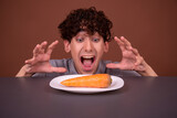 A funny young man looks at a plate from under the table. Diet and weight loss. Proper nutrition. Brown background. Copy the space.