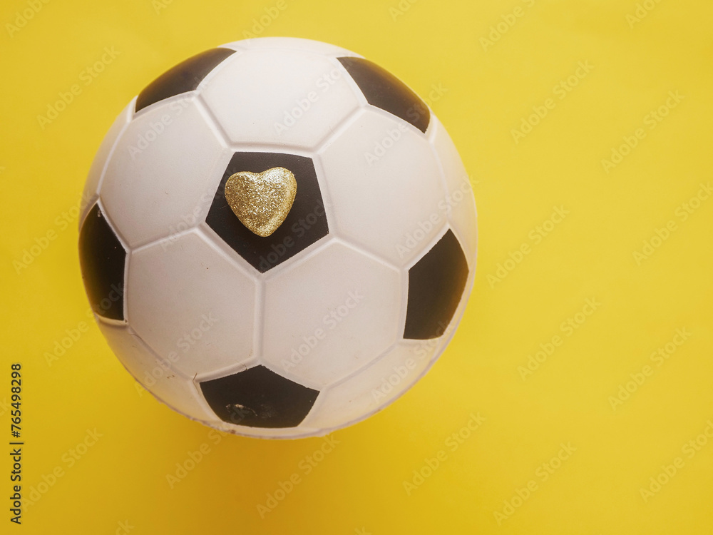 Small golden heart with glitter on classic old fashioned black and white soccer football ball on yellow background. Flat lay, Copy space for a message. Small golden heart with glitter on classic old fashioned black and white soccer football ball on yellow background. Flat lay, Copy space for a message.