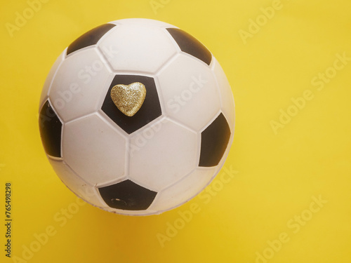 Small golden heart with glitter on classic old fashioned black and white soccer football ball on yellow background. Flat lay  Copy space for a message.