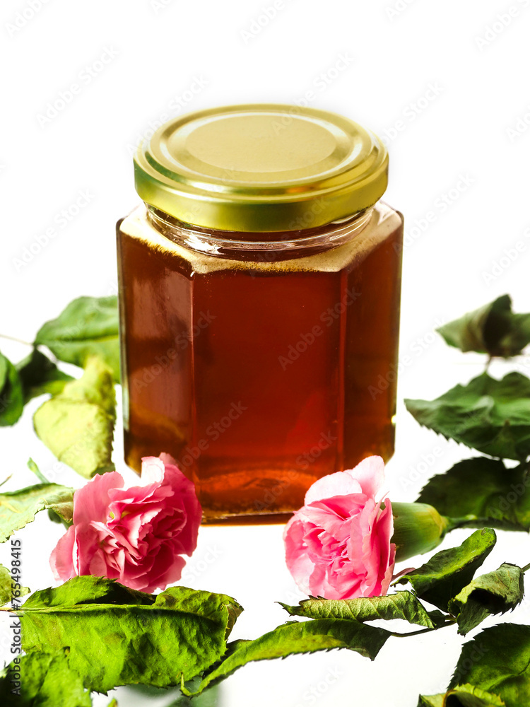 Glass bottle of fine honey with golden lid on white background with green leaf and red flowers. Agriculture organic product famous for its sweet taste and health benefits and value. Glass bottle of fine honey with golden lid on white background with green leaf and red flowers. Agriculture organic product famous for its sweet taste and health benefits and value.