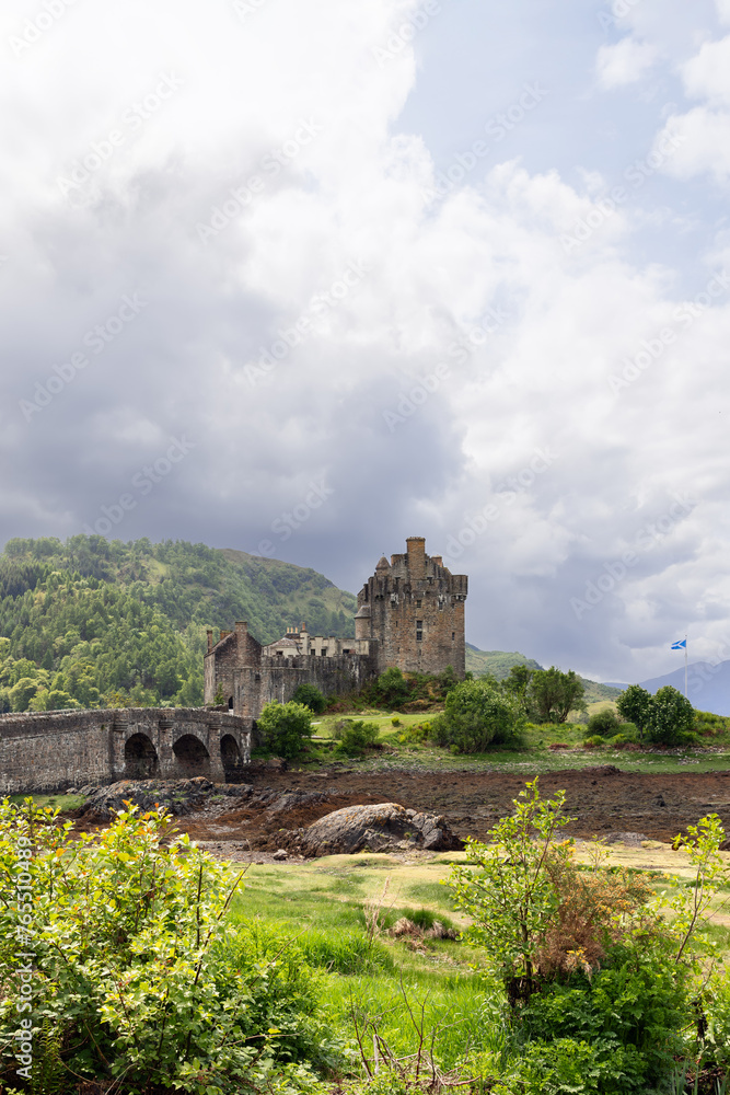 Clouds gather over the historic Eilean Donan Castle, where a stone bridge connects the iconic Scottish landmark to lush greenery, with the fluttering flag
