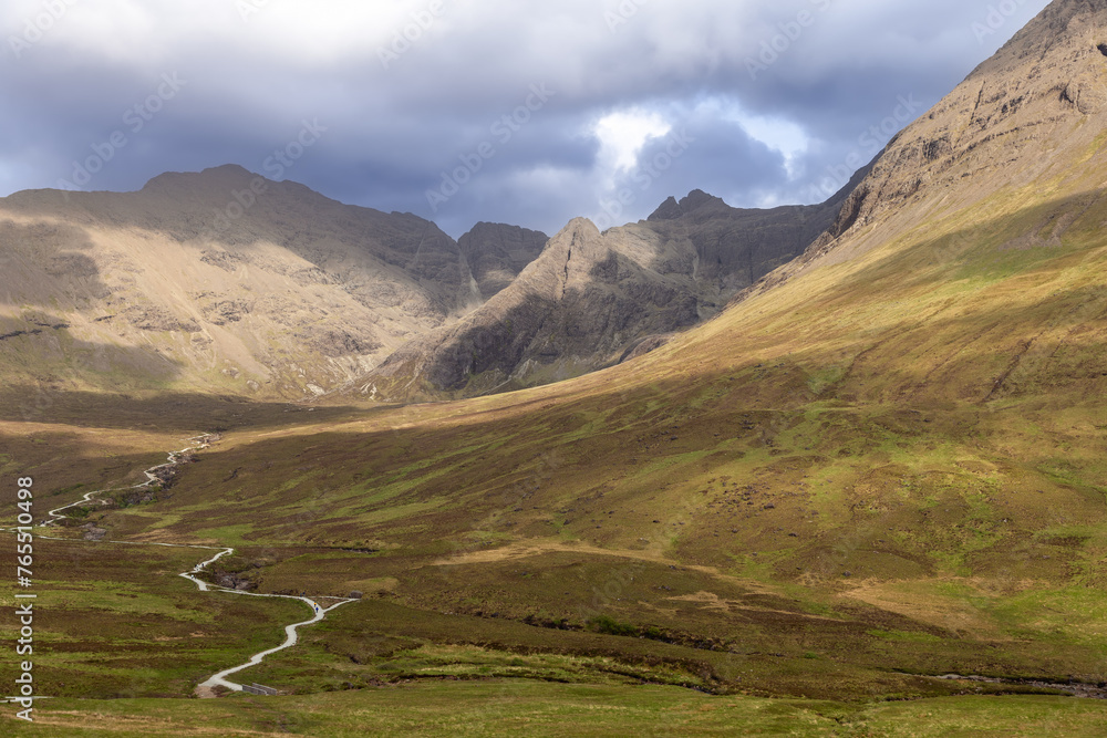 A meandering tourist path cuts through the verdant valley of the Isle of Skye, leading adventurers towards the Fairy Pools under a dramatic sky