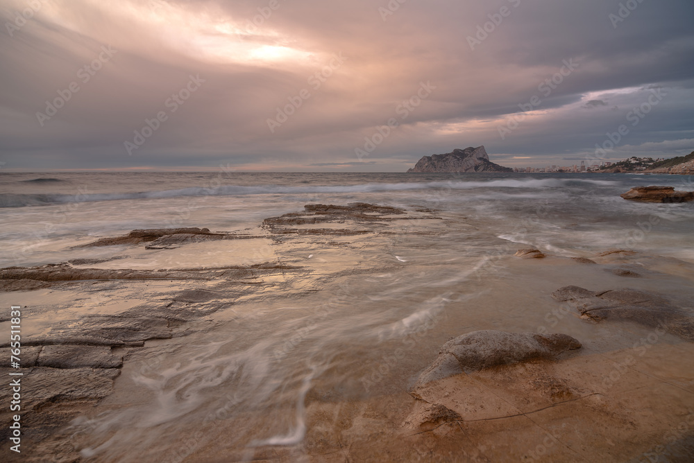 Coastal landscape with the Peñon de Ifach on the horizon. The sea is calm and the sky is cloudy.