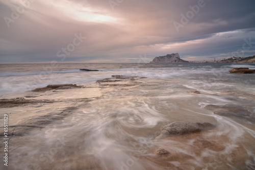 Coastal landscape with the Peñon de Ifach on the horizon. The sea is calm and the sky is cloudy.