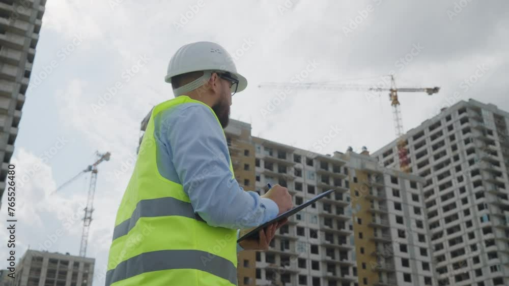 Bearded male construction supervisor in hard hat and reflective vest writing notes on clipboard with backdrop of high-rise buildings under construction and cranes Bearded male construction supervisor in hard hat and reflective vest writing notes on clipboard with backdrop of high-rise buildings under construction and cranes