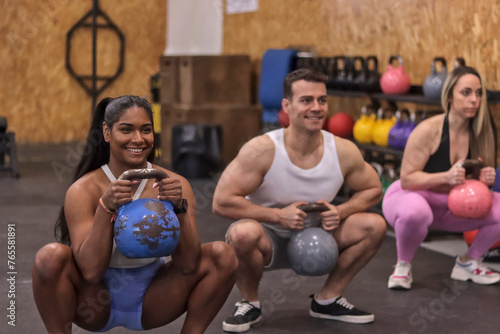 Group of multiracial smiling people doing squats with kettlebell dumbbells  in a row  inside a gym  sport dressed. Fitness concept