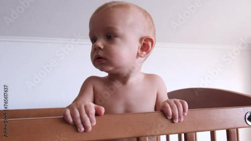 A child standing in a crib without a shirt holds on to the handrails and sadly calls his mother photo