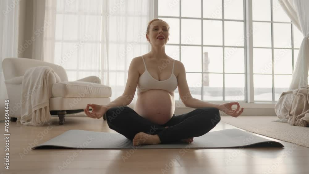 Pregnant woman doing meditation at home. Attractive female practices yoga stretching meditation, sitting at yoga mat at floor in lotus pose, deep breathing. Pregnant yoga, maternity sport concept.