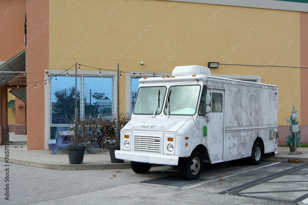 A white food truck, somewhat worn, is parked next to a yellow building with large windows; seems to be ready to start selling food.