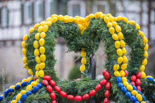 Osterbrunnen mit gelben, roten und blauen Ostereiern aus Kunststoff vor historischem Gebäude Pfarrhaus in Neckartenzlingen