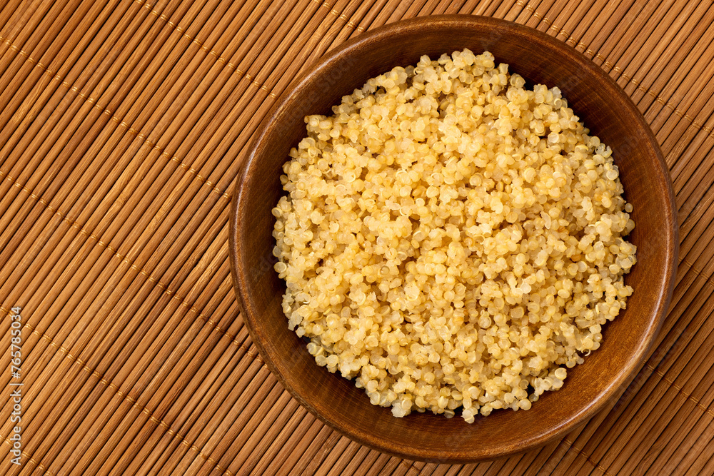 Cooked white quinoa in a dark wood bowl isolated on bamboo matt from above. Cooked white quinoa in a dark wood bowl isolated on bamboo matt from above.