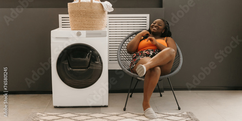 Happy plus size African woman waiting for clean clothes while sitting in chair near the washing machine in laundry